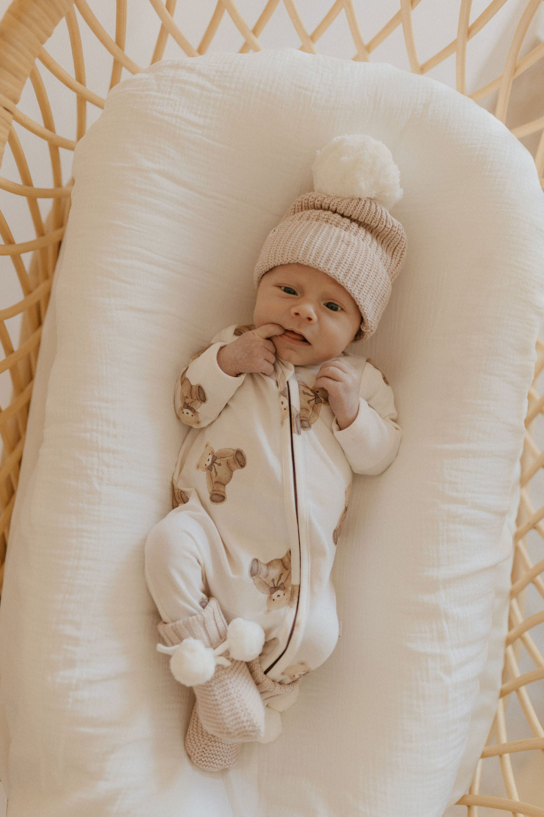 Baby wearing the Gingham Teddy Zipsuit and a knit beanie, lying in a bassinet